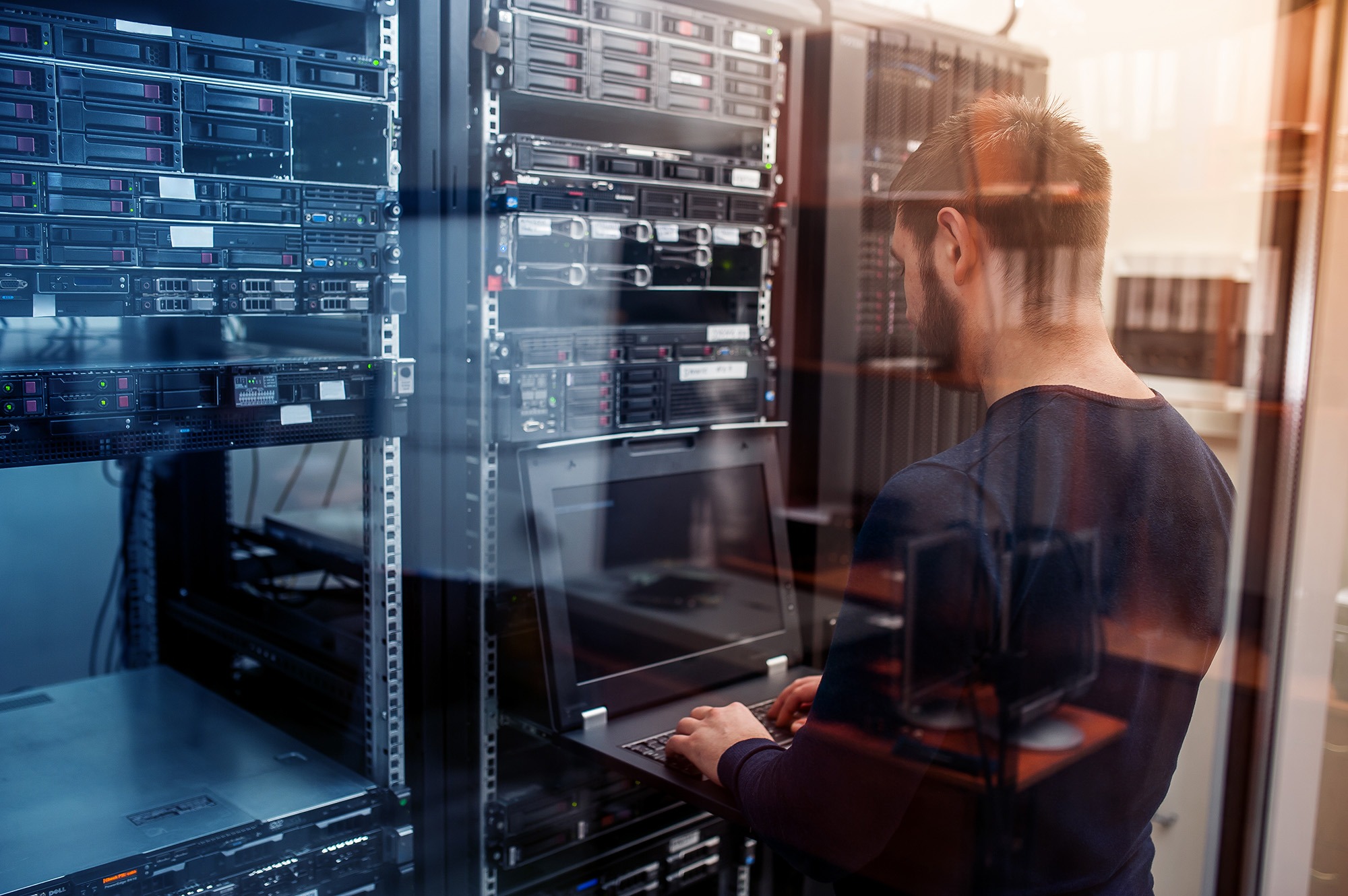 A technician works on a laptop in a server room, surrounded by stacked server racks, emphasizing the importance of IT infrastructure management.