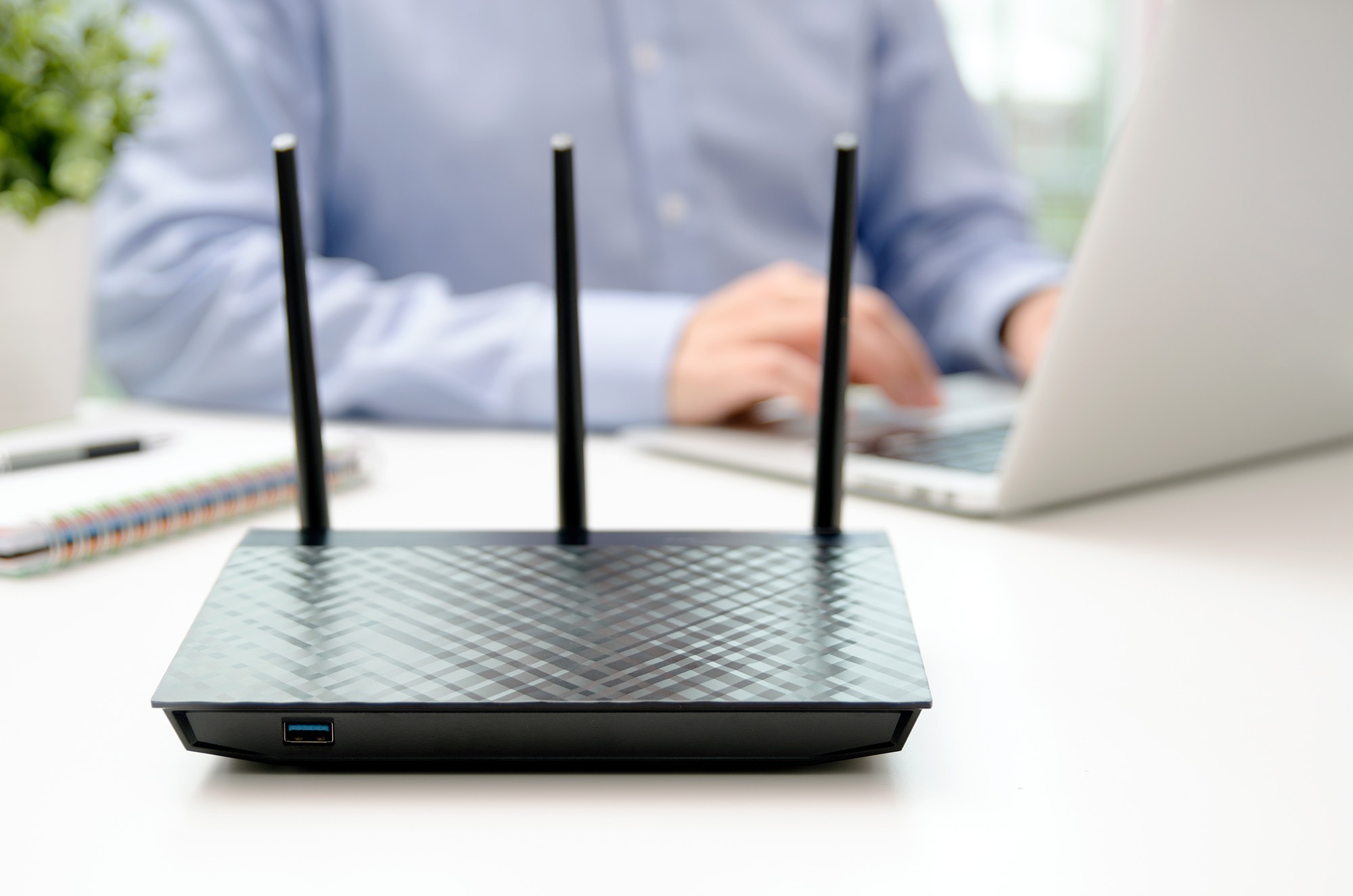 Wireless router with three antennas, positioned on a table. A person is using a laptop in the background, emphasizing home office connectivity.